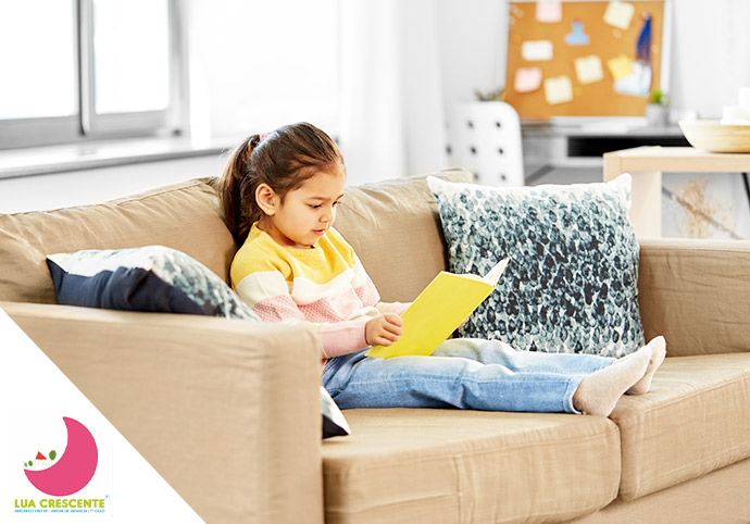 Menina sentada num sofá a ler um livro com conforto, apoiada numa almofada.